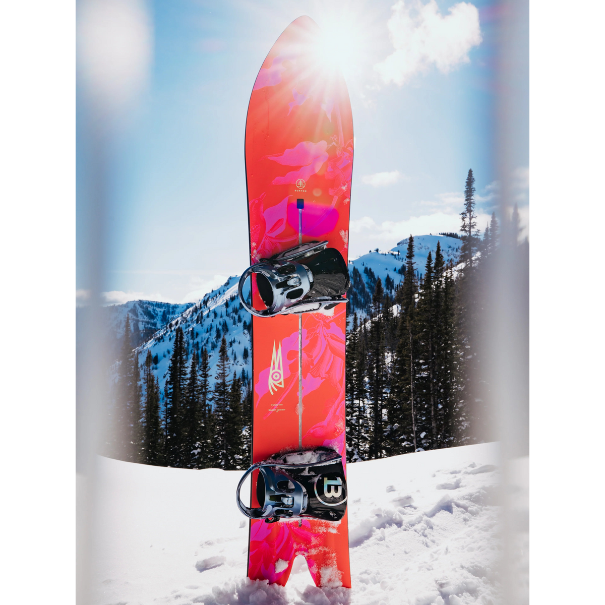 Burton Smooth Operator rose équipée de fixations, plantée verticalement dans la neige devant un paysage de montagne