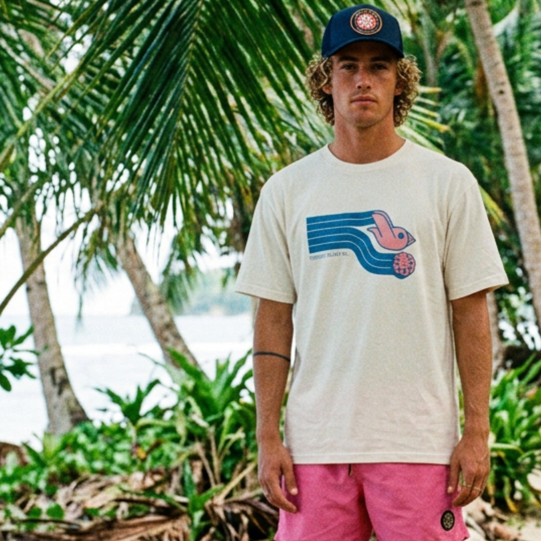 homme sur plage tropicale avec casquette bleue t-shirt blanc et boardshort surf rose