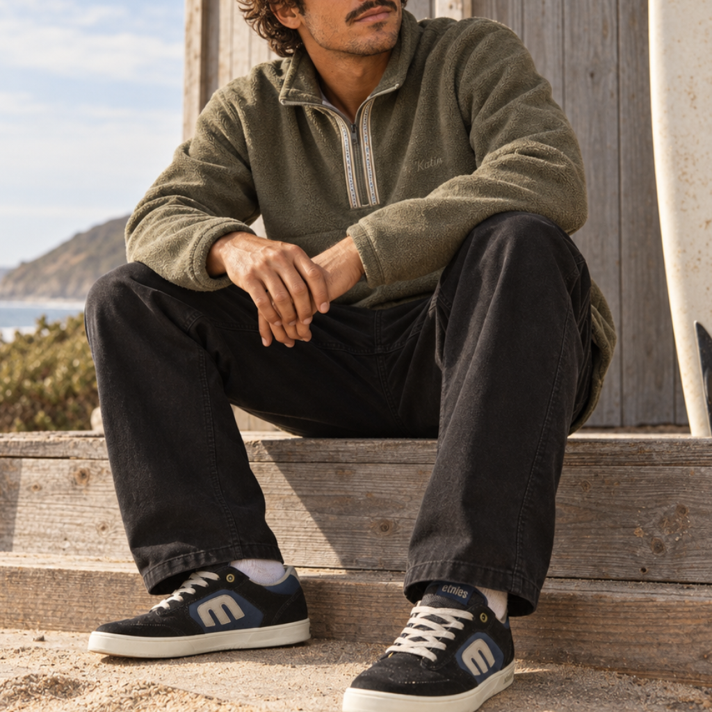 Pantalon chino Katin Clark noir porté par un homme assis sur des marches en bois, jambes droites, avec sweat vert et baskets etnies bleu marine.