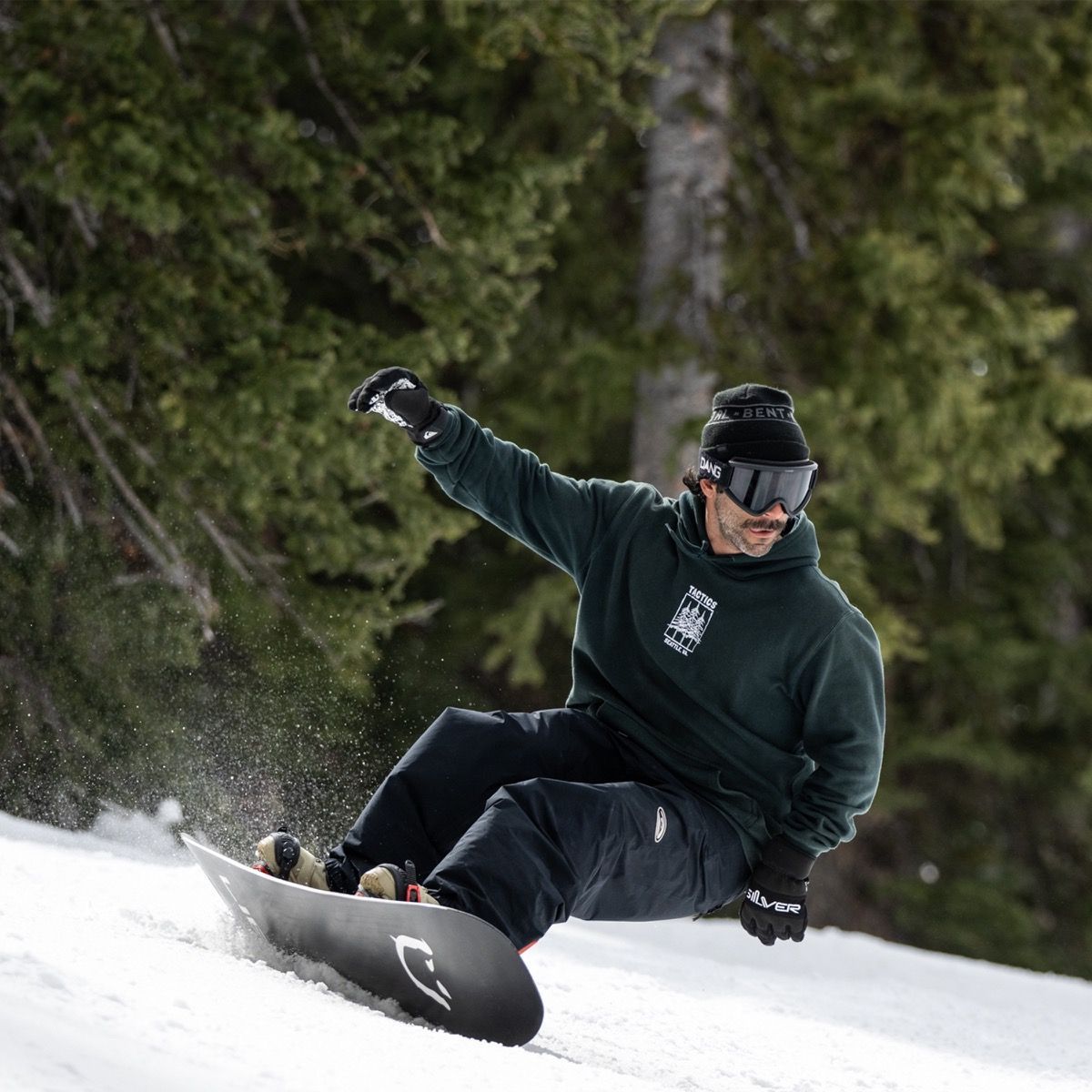 Snowboarder réalisant un virage coupé en appui sur la carre, planche inclinée et projection de neige sur piste bordée de conifères.