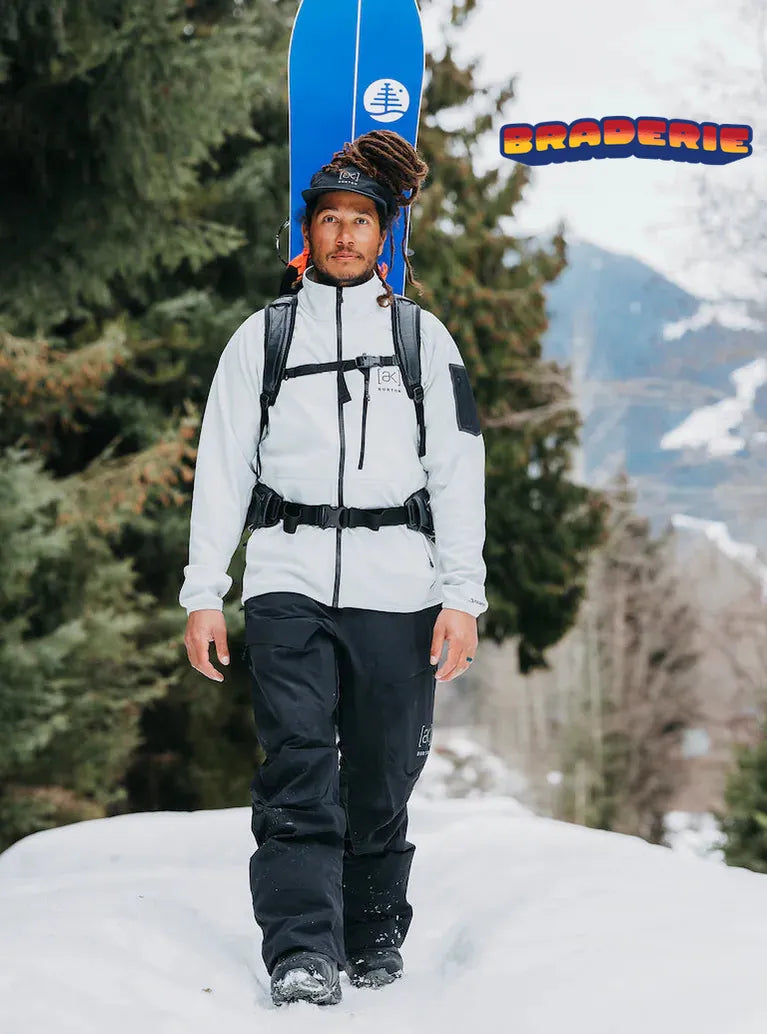 homme pantalon noir veste blanche de ski et snowboard dans le dos lifestyle