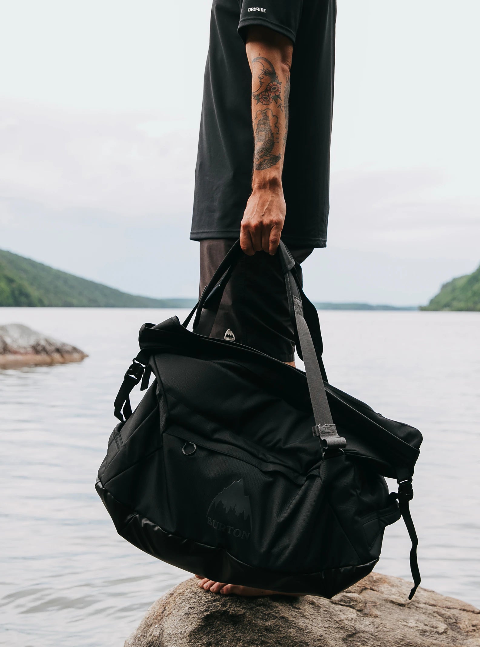 Sac Burton Multipath Duffel 60L Black porté à la main par un homme arriere plan lac