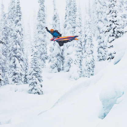 Snowboarder en plein saut dans une forêt enneigée, semelle orange et noire du Burton Smooth Operator visible sous la planche