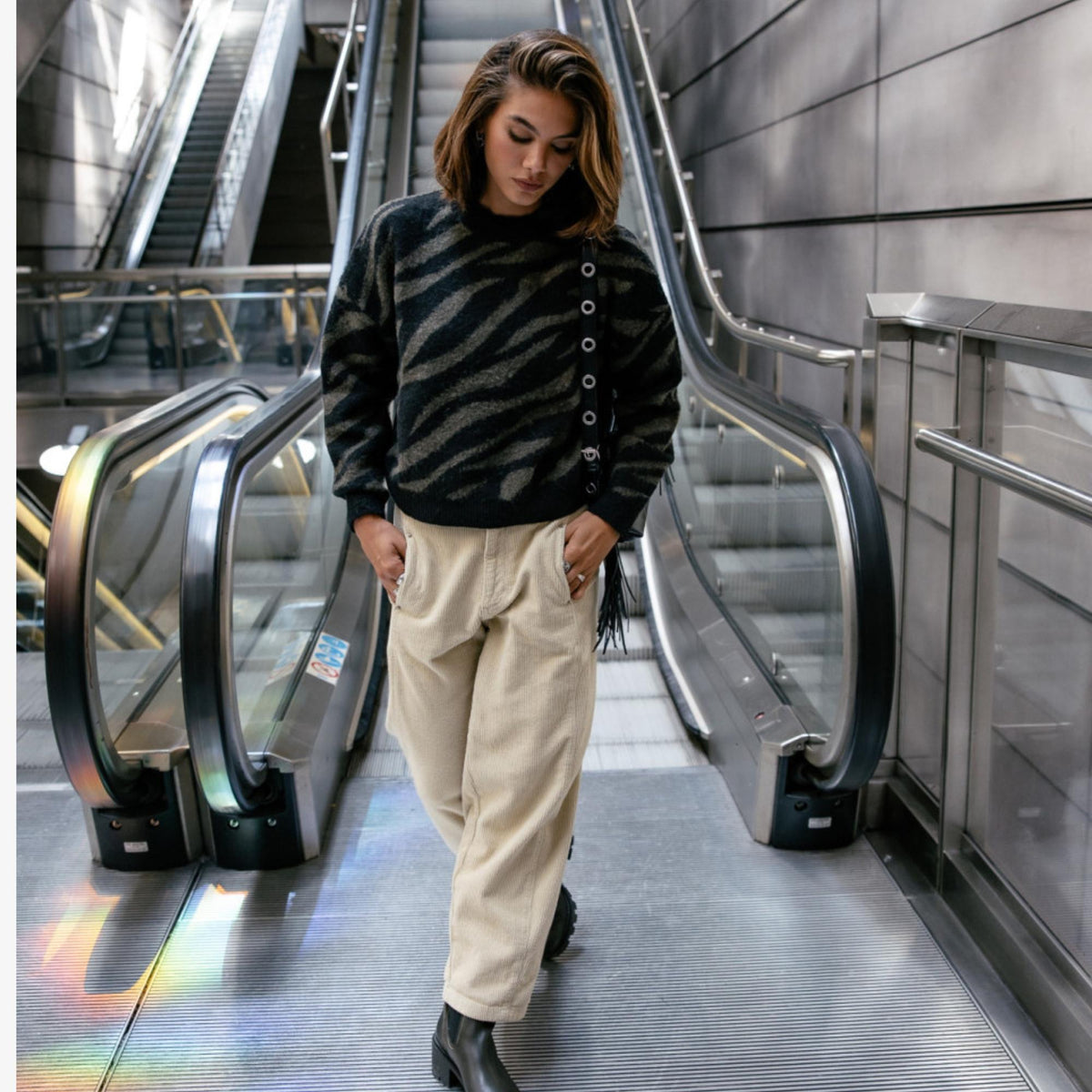 Look féminin avec pantalon Rosalia Veluto beige crème, pull rayé sombre et chaussures noires dans un escalator.