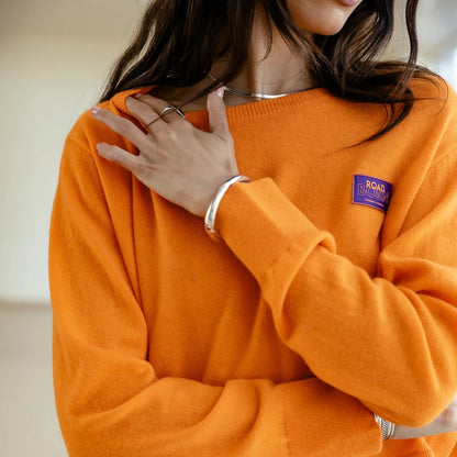 Détail du pull orange avec poignets longs resserrés et patch poitrine violet brodé de lettrage contrastant