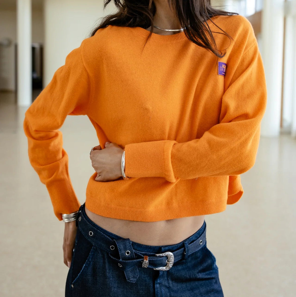 Rendu porté de face du pull court orange, épaules tombantes et manches longues structurées par des poignets ajustés