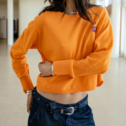 Rendu porté de face du pull court orange, épaules tombantes et manches longues structurées par des poignets ajustés