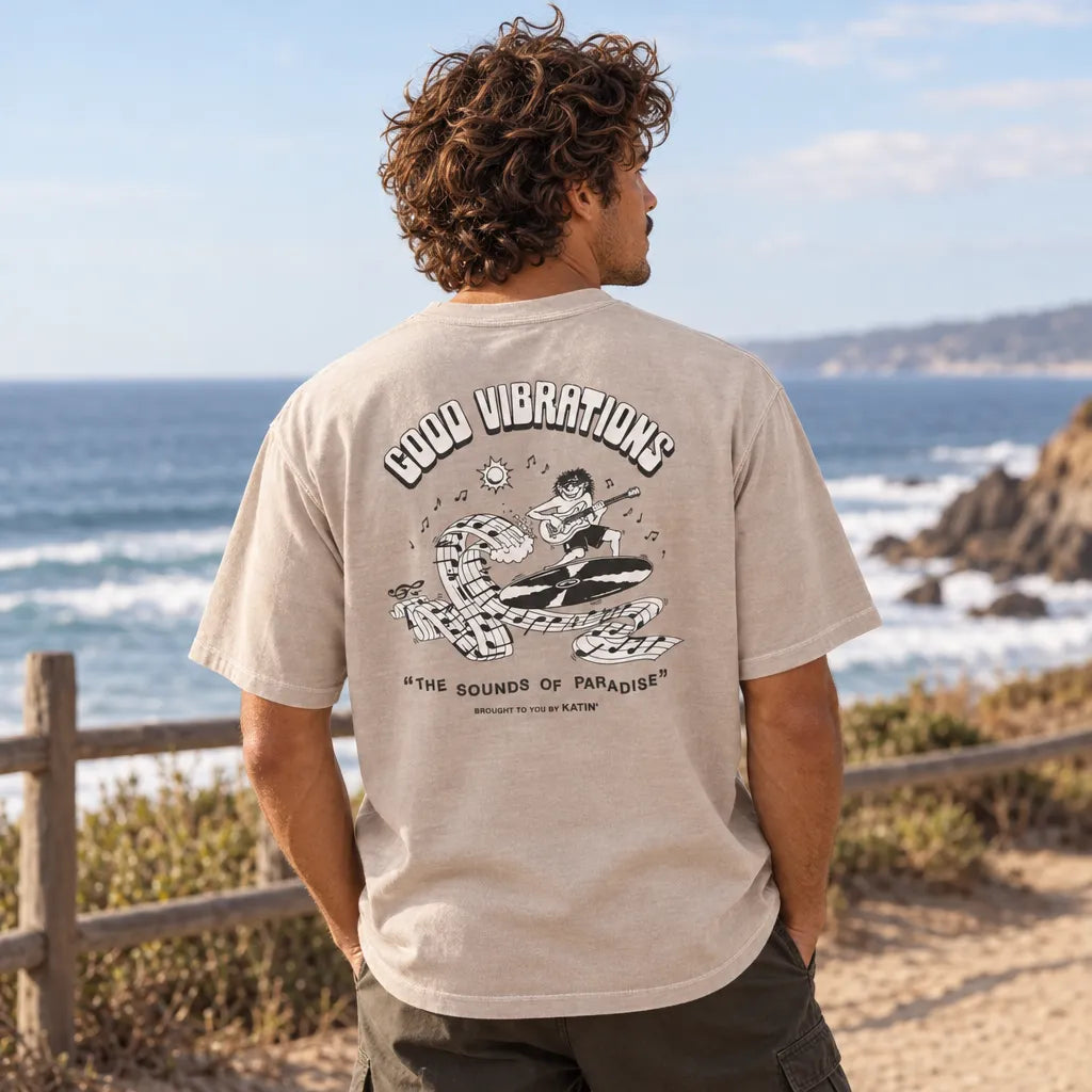 Homme portant de dos un t-shirt Katin Surf Rock beige clair, grand imprimé circulaire avec lettrage et illustration de vagues créées avec note de musique, face à l'océan.