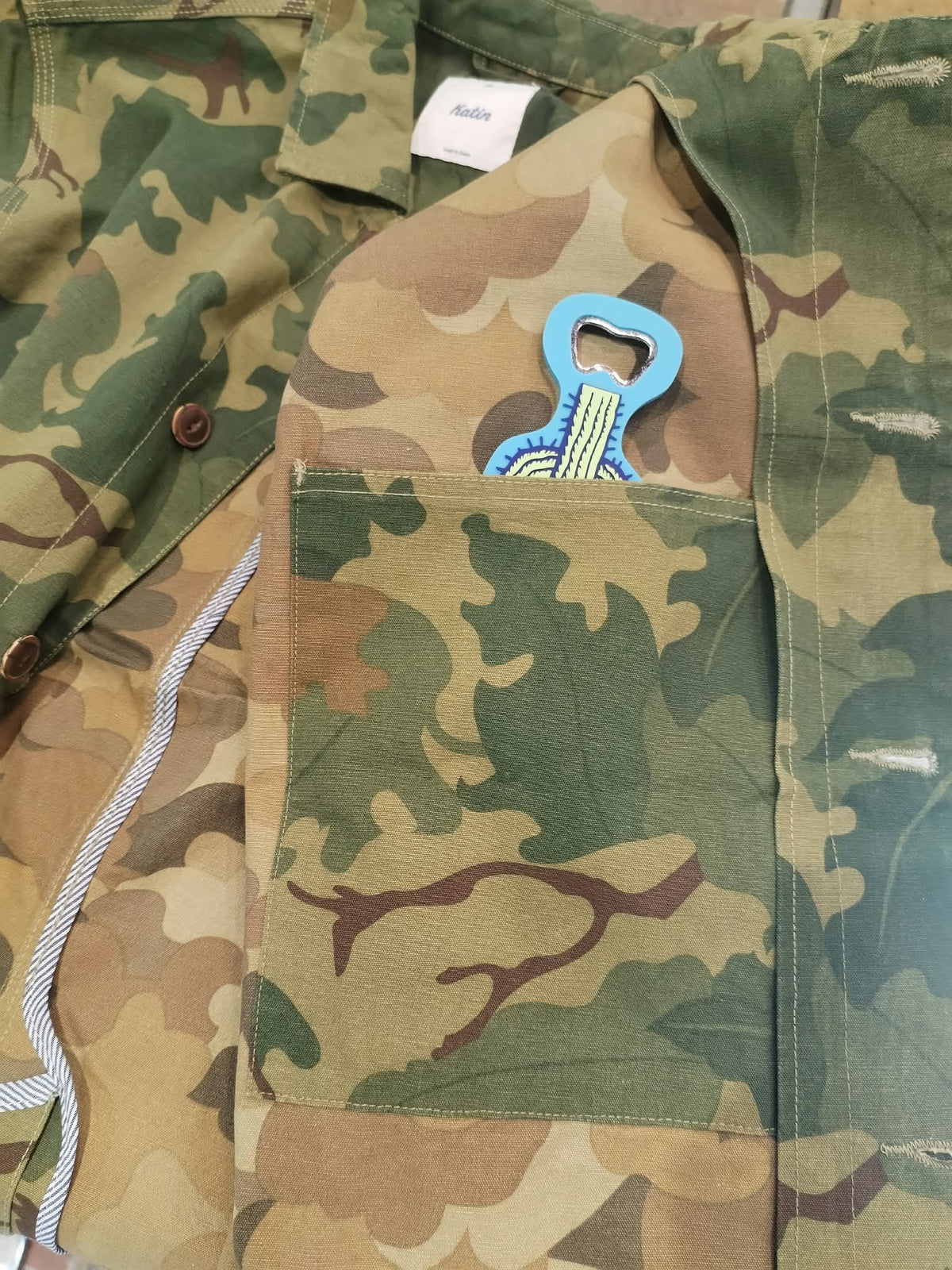 Détail intérieur de la veste Katin Task Coat, tissu camouflage, grande poche plaquée et étiquette blanche près du col.
