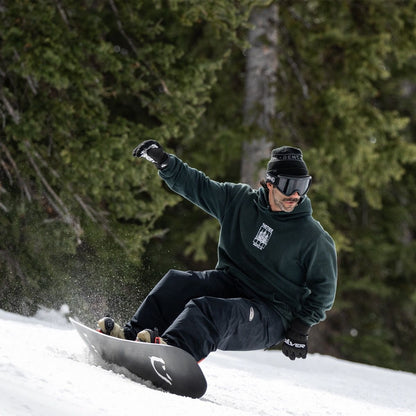 Snowboarder réalisant un virage coupé en appui sur la carre, planche inclinée et projection de neige sur piste bordée de conifères.