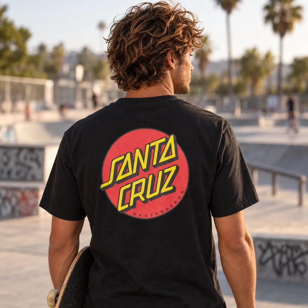 Homme portant le tee-shirt Santa Cruz Classic Dot noir, vue de dos avec skateboard à la main dans un skatepark au coucher du soleil.
