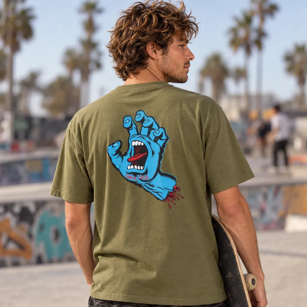 Skateur portant le tee-shirt Santa Cruz Screaming Hand Deep Olive, vue de dos avec skateboard sous le bras dans un skatepark bordé de palmiers
