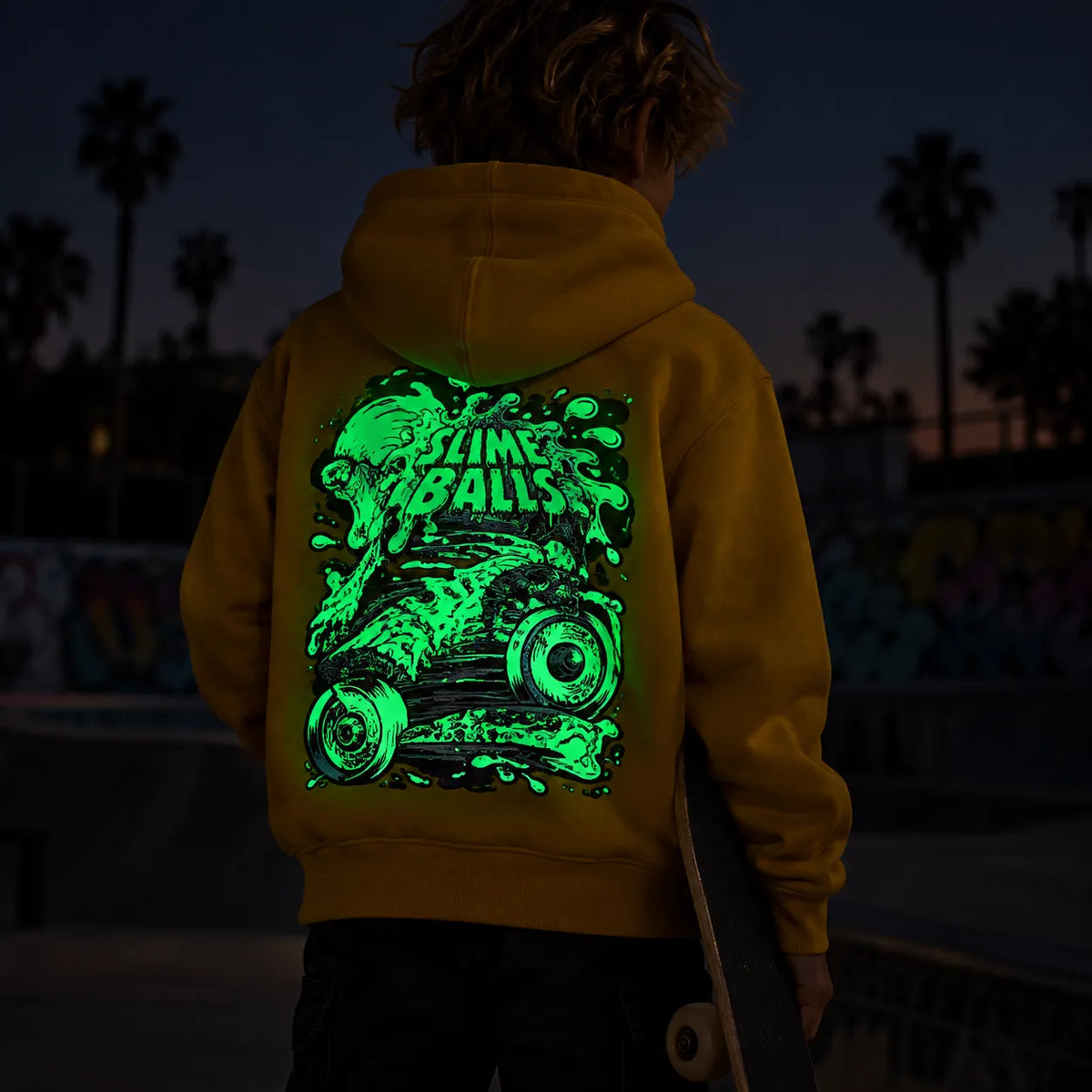 Enfant de dos au skatepark de nuit portant le hoodie Santa Cruz Youth Slime Balls Accelerator Mustard avec grand graphisme Slime Balls phosphorescent vert dans le dos